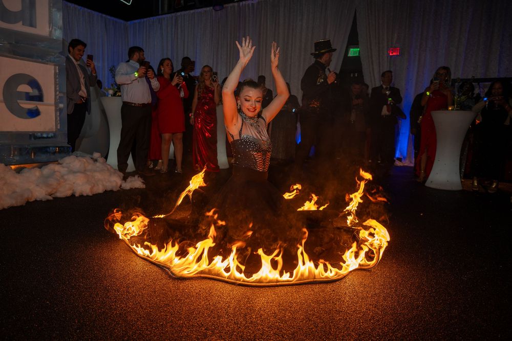 A performer dances energetically inside a ring of fire while spectators watch from the background in a dark venue.