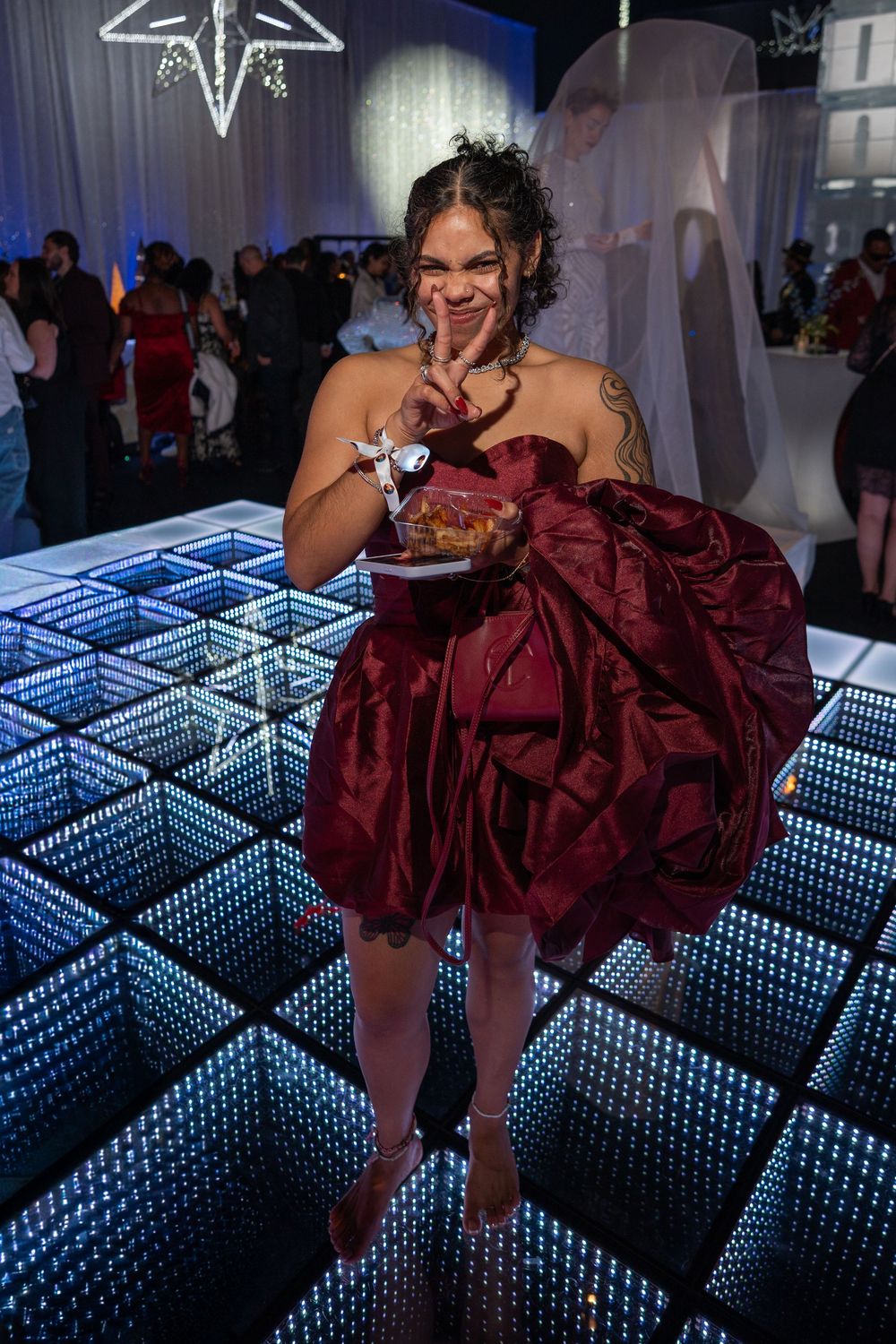 A person in a burgundy strapless dress enjoys food while standing on an illuminated LED dance floor at an event.