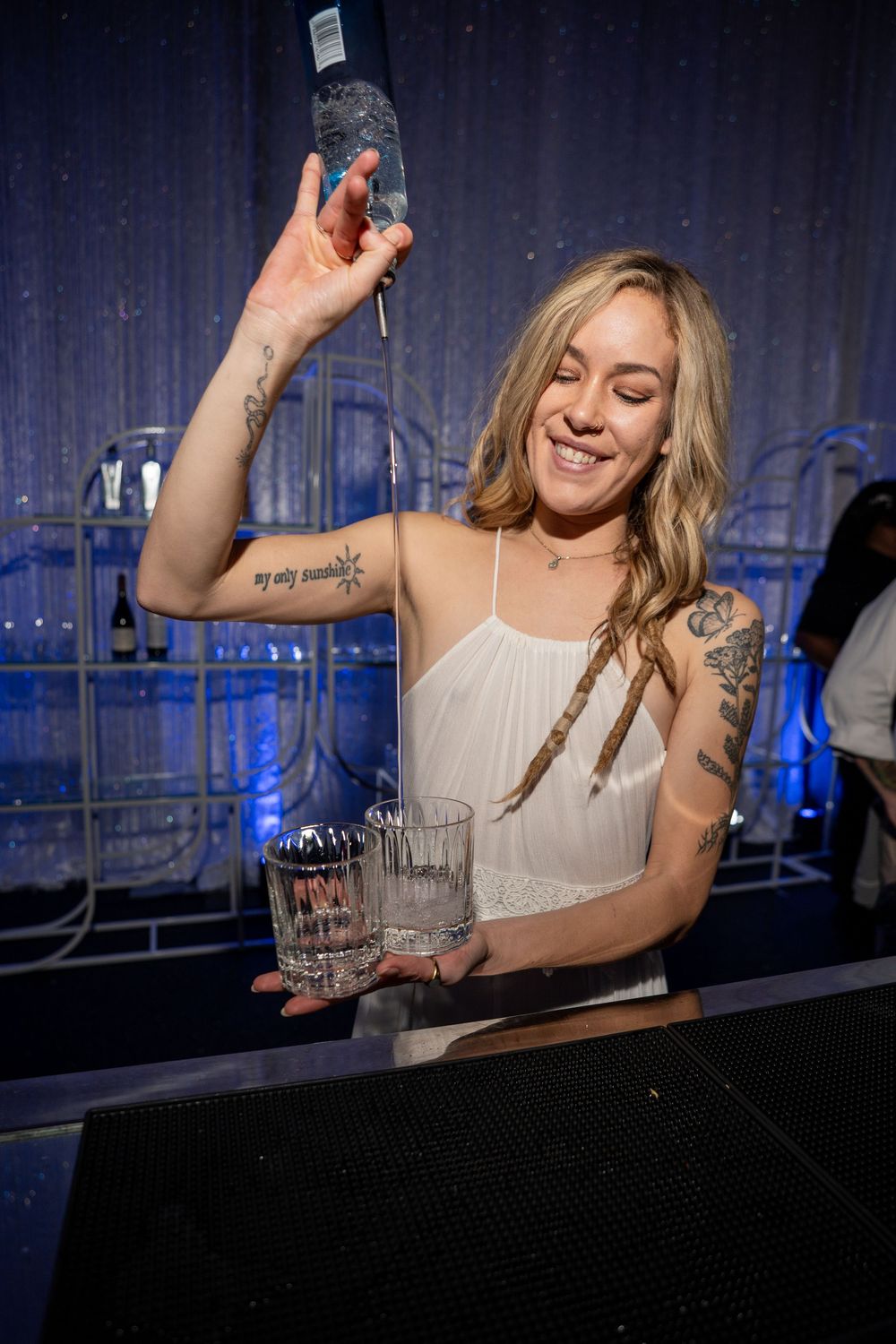 Bartender in white tank top pours drinks at a dimly lit bar with blue lighting in the background.