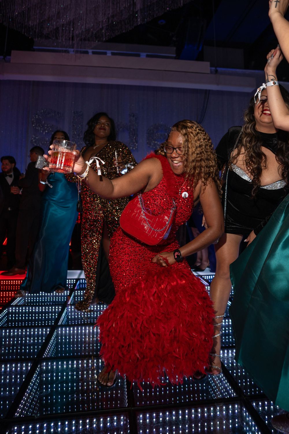 People dancing and celebrating on an illuminated dance floor at a party, with someone in a vibrant red dress in focus.
