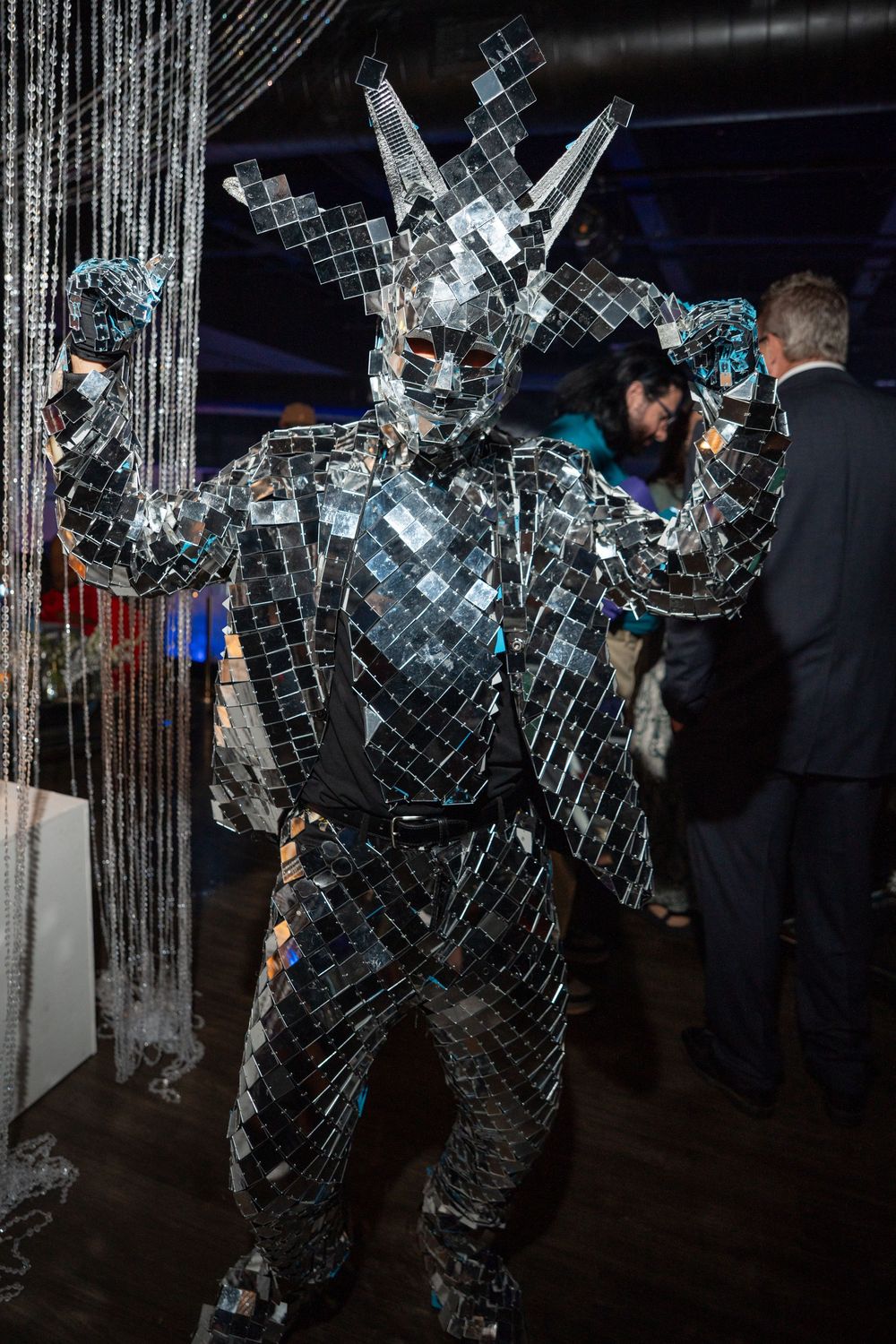 A person in a mirror ball disco suit dances and poses in a dark nightclub setting with reflective lighting effects.