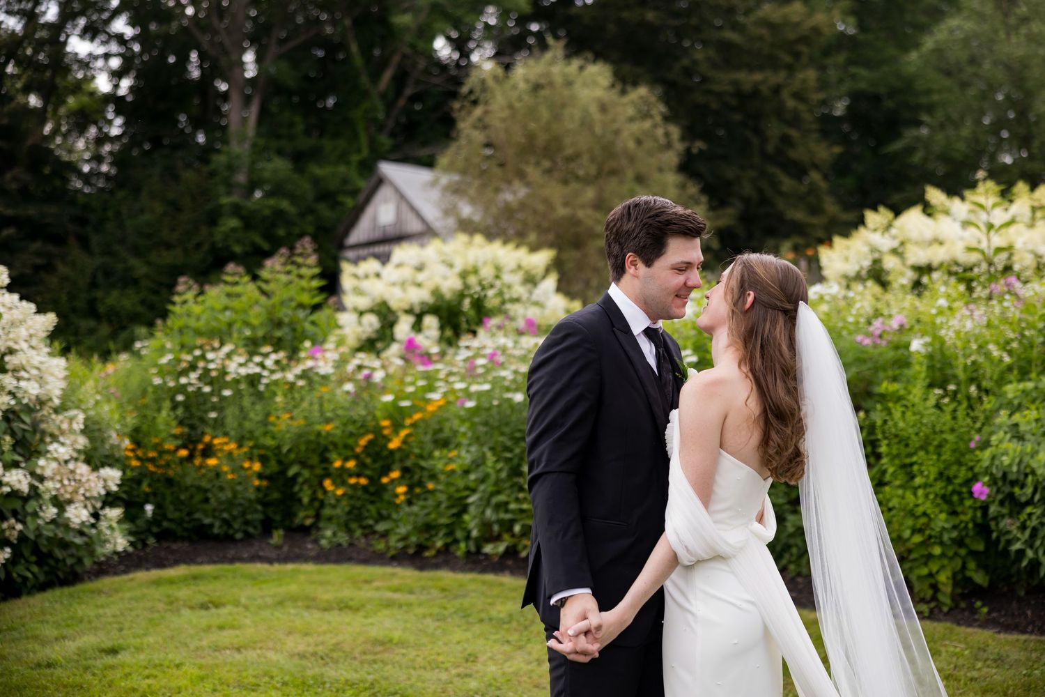 A romantic wedding couple stands together in a lush garden filled with colorful summer flowers and greenery.