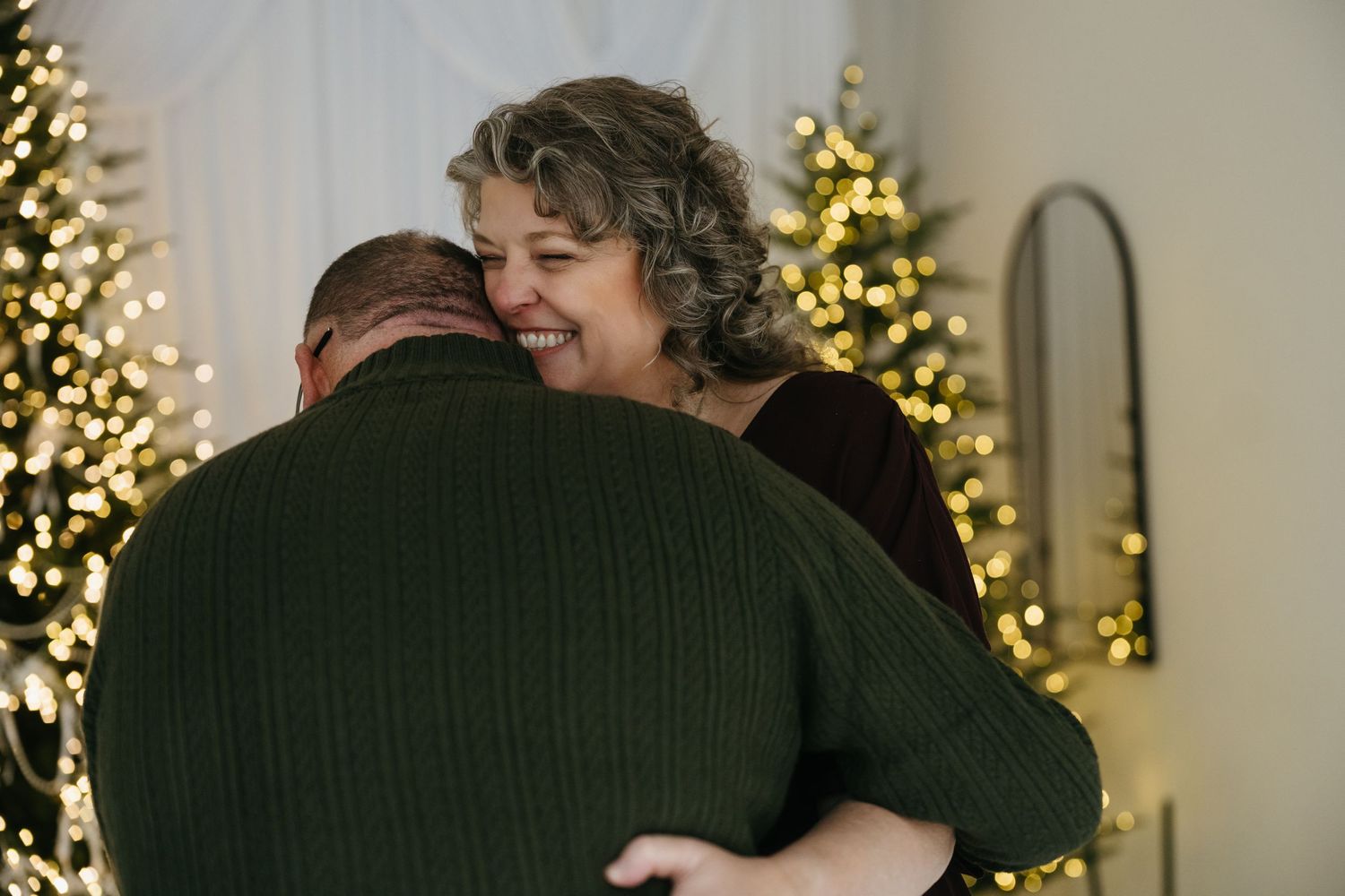 A couple shares a warm embrace during the holidays with Christmas lights twinkling in the background.