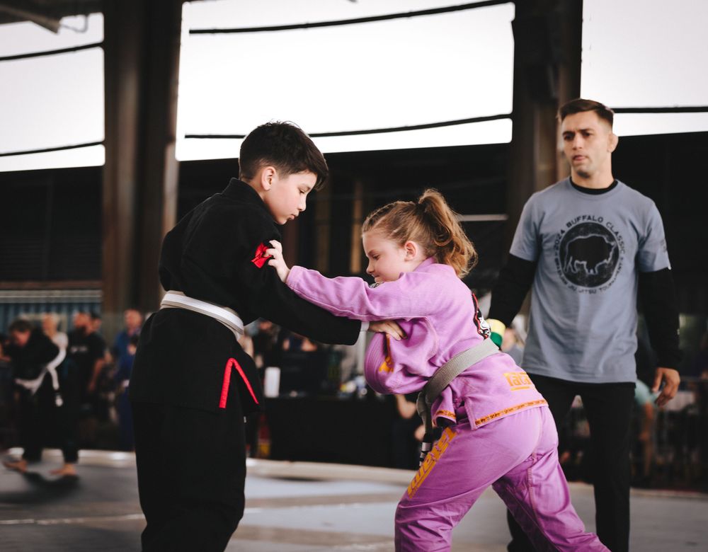 Two children practicing martial arts moves while instructor watches in background.