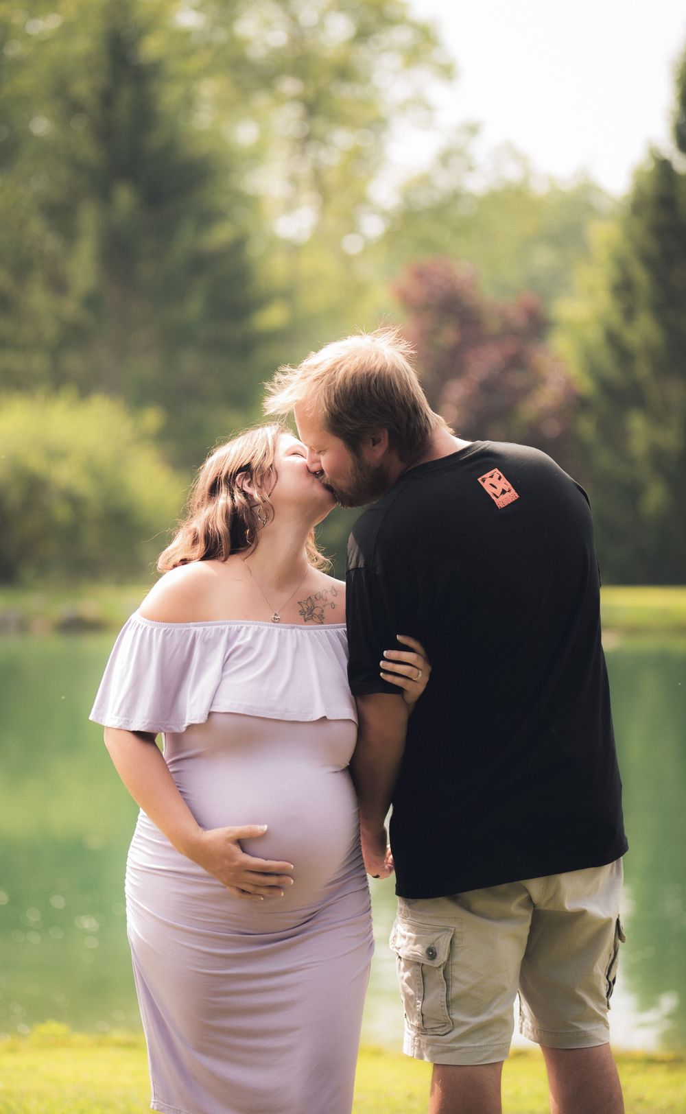 Expecting couple shares an intimate moment outdoors in casual summer attire.
