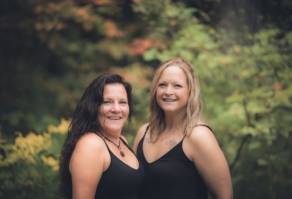 Two women in black tops smile together outdoors against a blurred natural background.