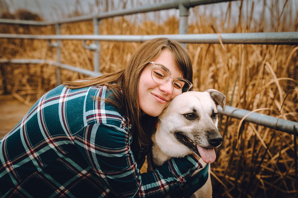 A happy white dog and someone in a plaid shirt share an affectionate moment among tall reeds.