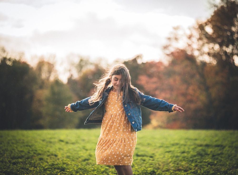 Person in denim jacket and yellow dress twirls joyfully in an autumn field at sunset.