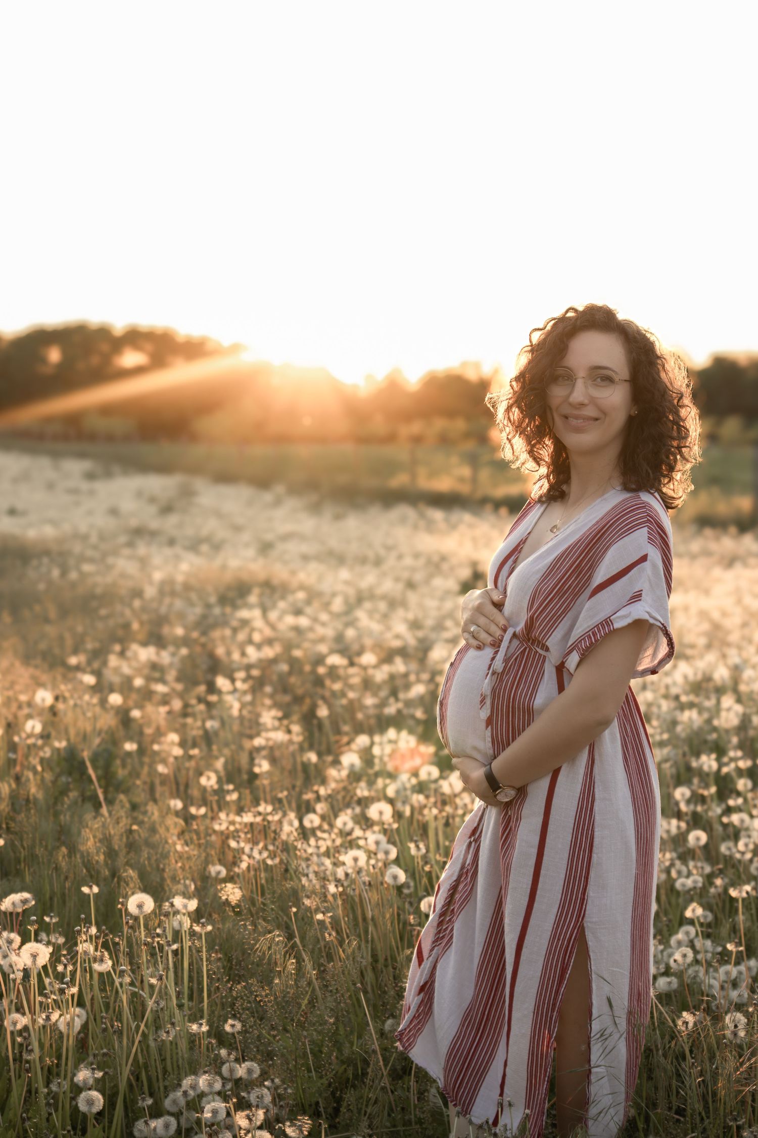 Maternity photo of woman in striped dress standing in field of wildflowers at sunset.