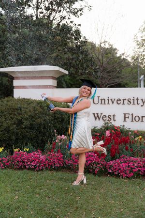A graduate in a white dress poses playfully by a university sign surrounded by vibrant pink flowers and greenery.