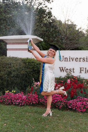 A cheerleader performs a high kick pose in front of a university sign surrounded by vibrant pink flowers.