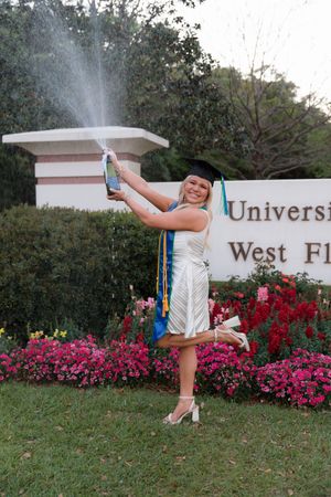 A graduate in white dress kicks up their leg playfully while posing with their diploma in front of blooming azalea bushes.