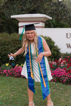Graduate in blue and gold stole celebrates on campus lawn with pink flowers in background.