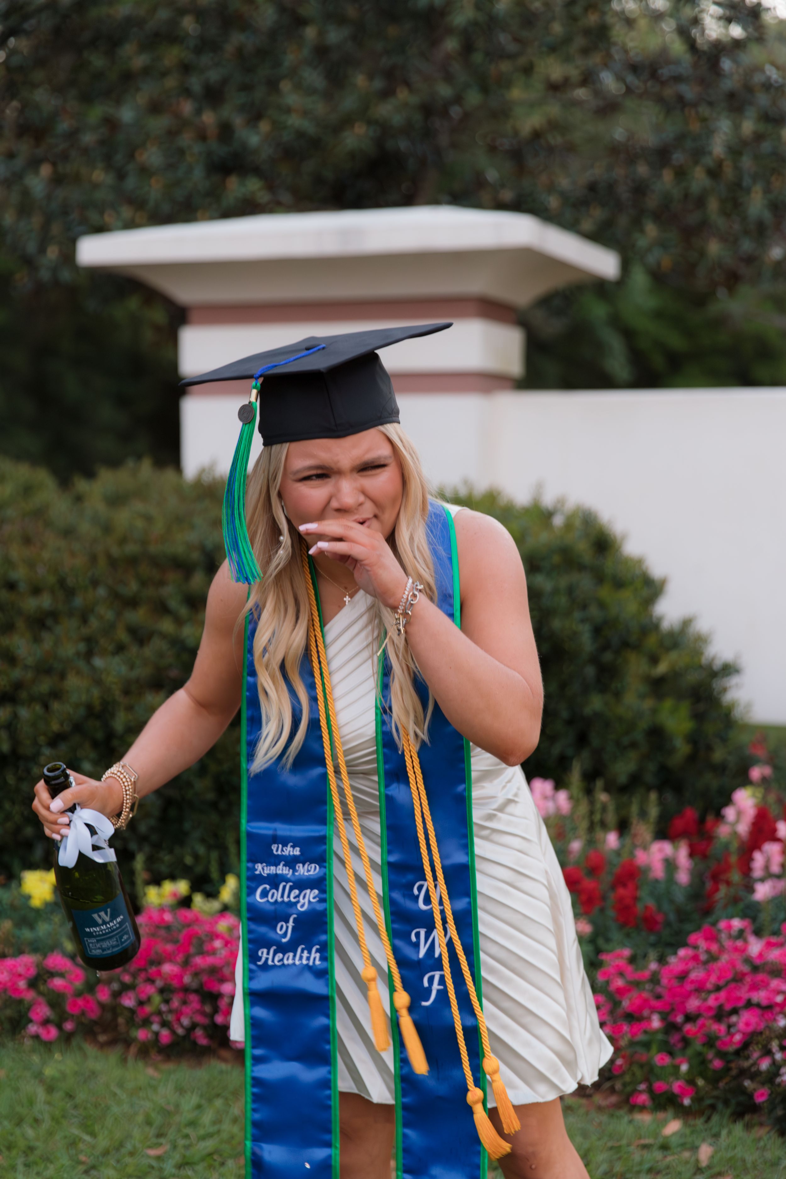 Graduate in cap and gown with graduation stole and sash poses joyfully in garden with pink flowers.
