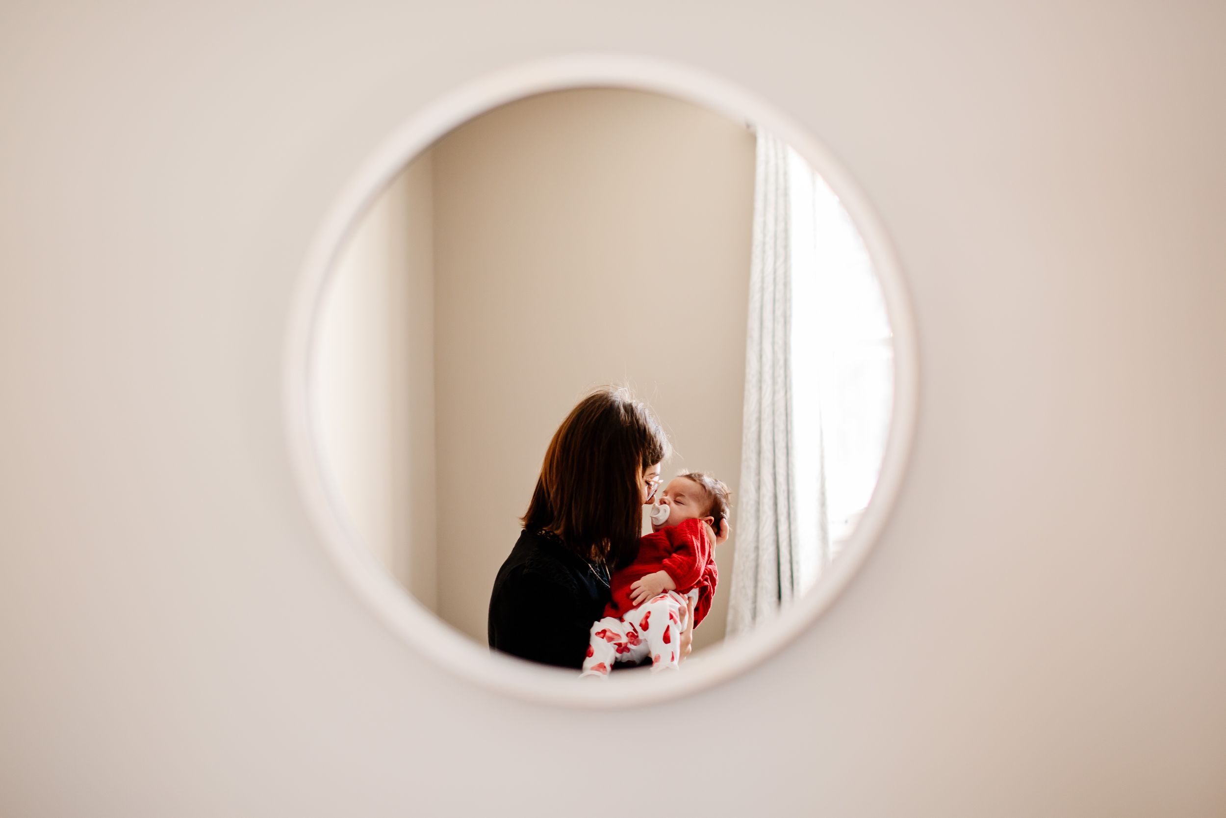 Mother holding newborn baby reflected in a round white-framed wall mirror.