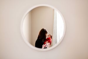 Mother holding newborn baby reflected in a round white-framed wall mirror.