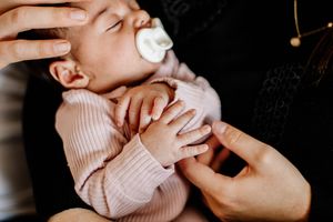 Sleeping newborn baby in pink outfit with pacifier having tiny hands held by parent.