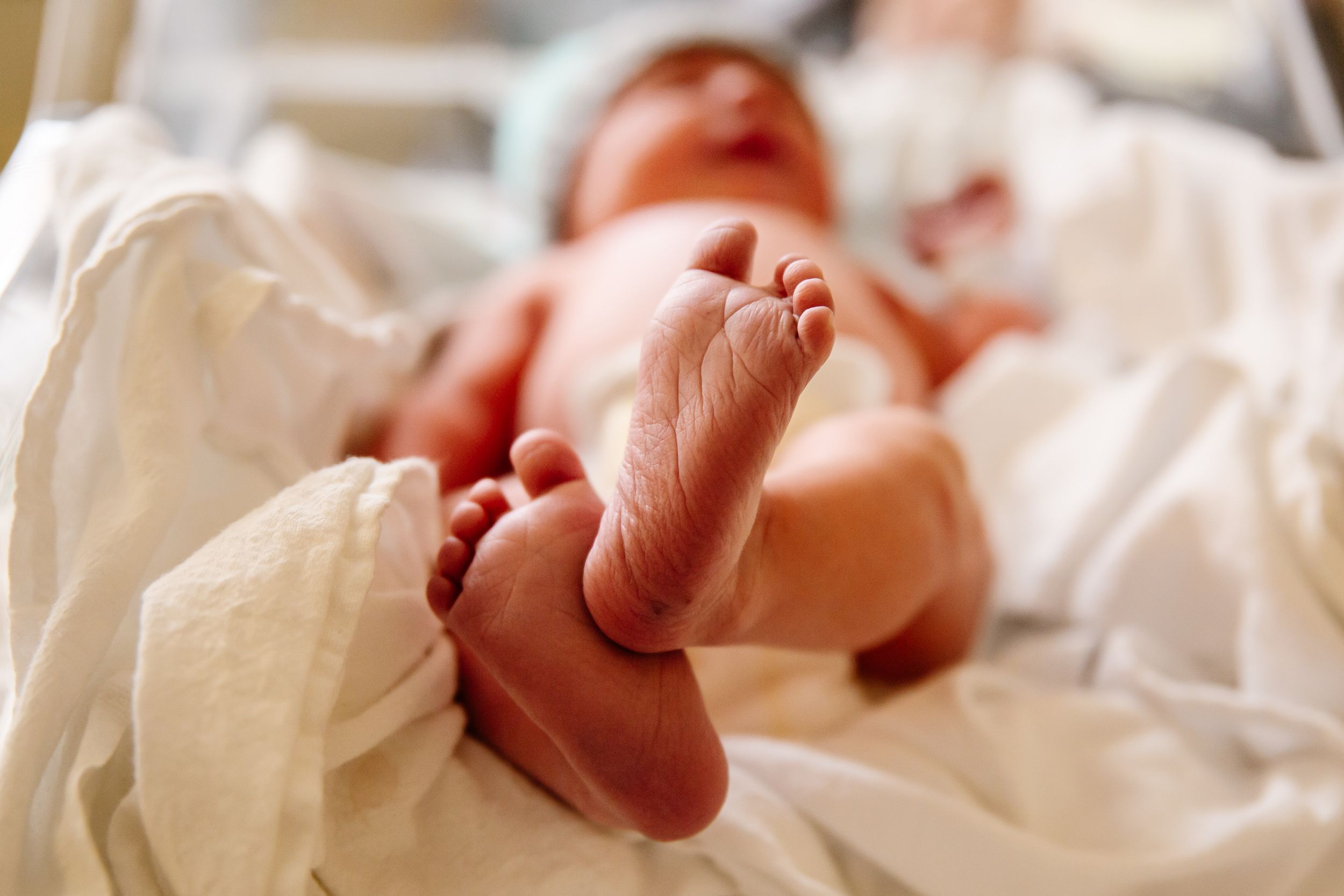 Newborn baby lying on white hospital blankets with tiny feet raised in the air.