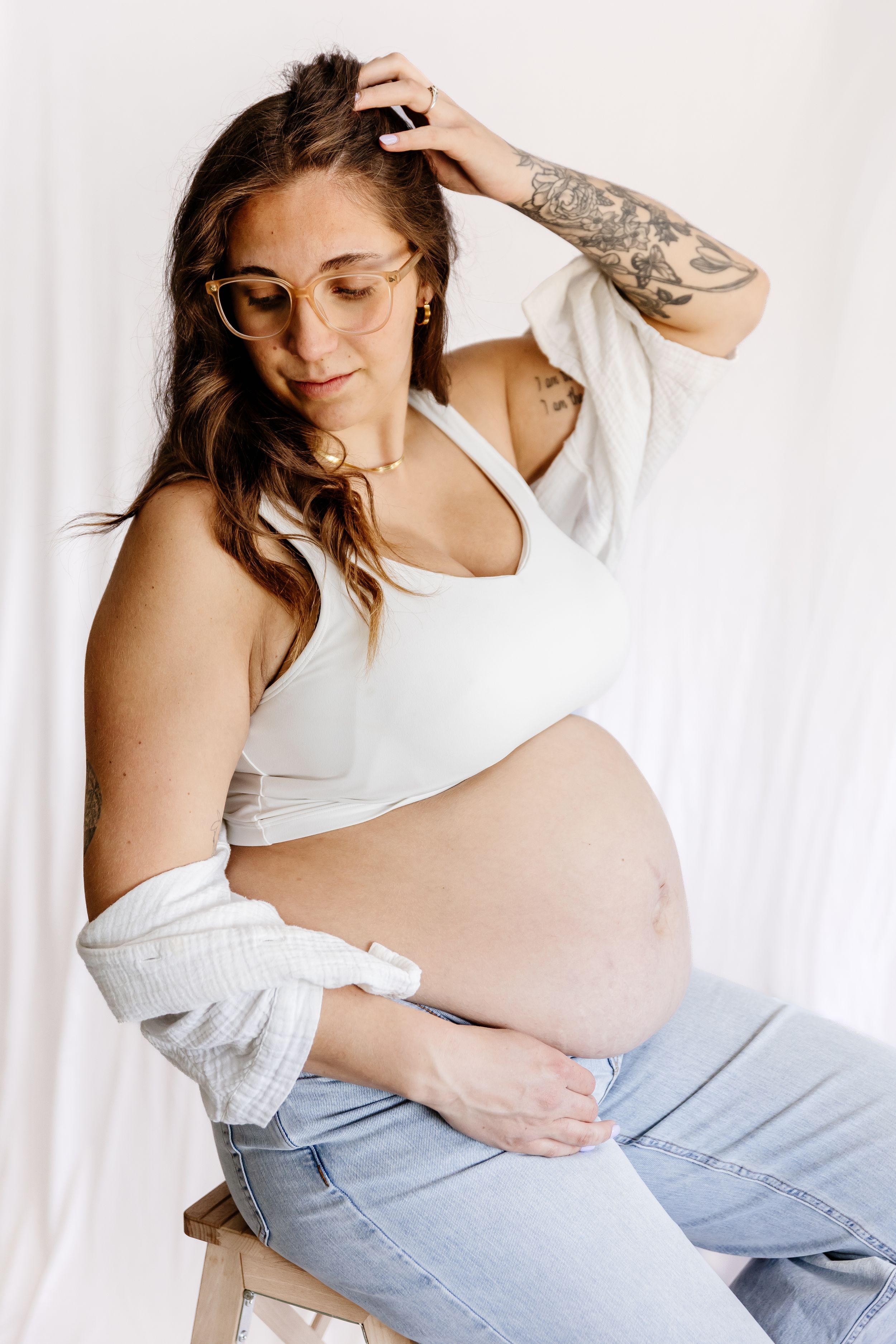 Pregnant woman with tattoos sits on stool in white crop top and jeans, cradling her baby bump in a bright studio setting.