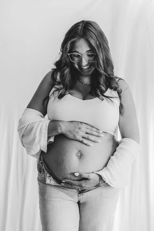 Black and white photo of smiling pregnant woman in glasses cradling her baby bump, wearing crop top and jeans.