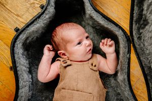 Newborn baby in brown romper lying inside an open guitar case on a wooden floor.