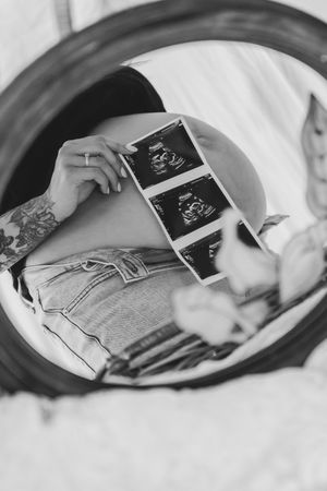 Black and white photo of pregnant woman holding ultrasound scans reflected in round mirror, wearing jeans with tattoo visible.