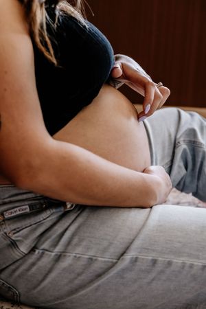 Pregnant woman in black crop top and unbuttoned jeans cradling her bare baby bump with both hands.