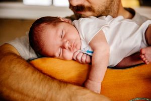 Sleeping newborn baby resting peacefully on father's chest beside an acoustic guitar.