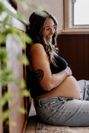 Smiling pregnant woman with glasses and tattoo sits against wall cradling baby bump, wearing crop top and jeans.