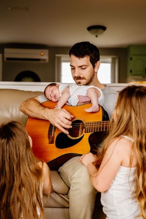 Father playing acoustic guitar with sleeping newborn on his chest surrounded by children.