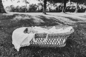 Black and white photo of a sleeping newborn baby in a wicker Moses basket on grass outdoors.