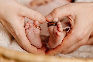 Parents with wedding rings gently cradle their newborn baby's tiny feet in a loving heart shape gesture.