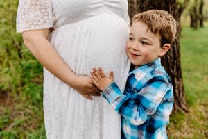 Young boy in plaid shirt hugs pregnant mothers belly outdoors near tree.