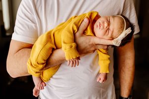 A man in a white t-shirt cradles a sleeping newborn baby dressed in a mustard yellow outfit and bow headband.