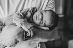 Black and white photo of a sleeping newborn baby cradled in an adult's arms, wearing a striped onesie.
