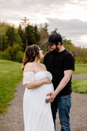 Expecting couple shares tender moment on garden path at sunset, woman in white lace dress.