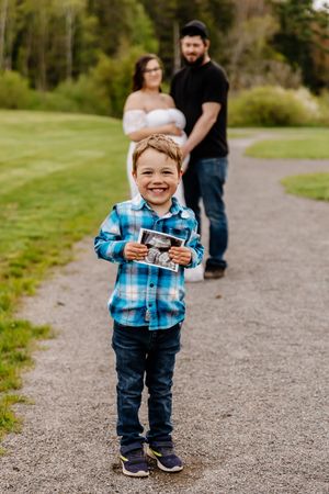 Smiling boy holds ultrasound photo on gravel path with pregnant mother and father standing behind him.