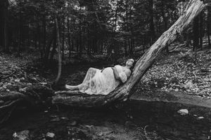 Black and white photo of pregnant woman in white gown resting on fallen log in forest.
