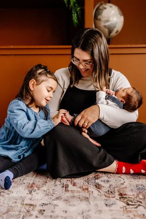 A smiling mother sits on a rug holding a newborn while her young daughter gently touches the baby's hand.