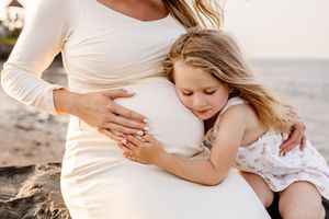 Young girl hugging her pregnant mother's belly on a beach at sunset.