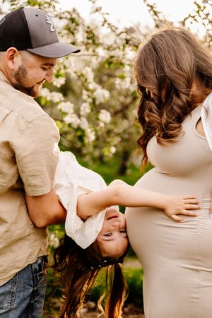 A happy family maternity photo with dad holding toddler upside down beside pregnant mom in a blooming orchard.