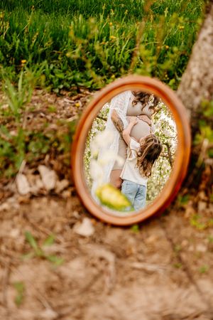 Oval mirror reflecting a couple embracing outdoors, leaning against a tree with green grass in the background.