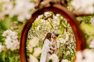 Couple embracing in a blooming spring orchard, framed by a vintage oval mirror surrounded by white flowers.