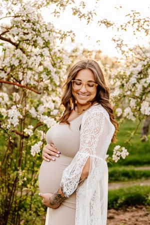 Smiling pregnant woman in beige dress and white lace cardigan cradling baby bump among blooming white spring trees.