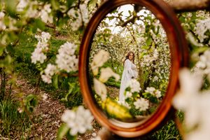 Woman in white dress reflected in oval mirror surrounded by blooming white apple trees in lush green garden.