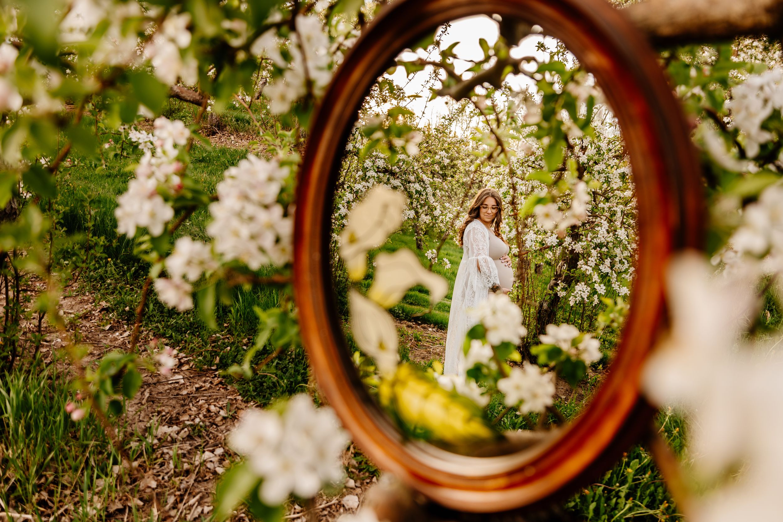 Woman in white dress reflected in oval mirror surrounded by blooming white apple trees in lush green garden.