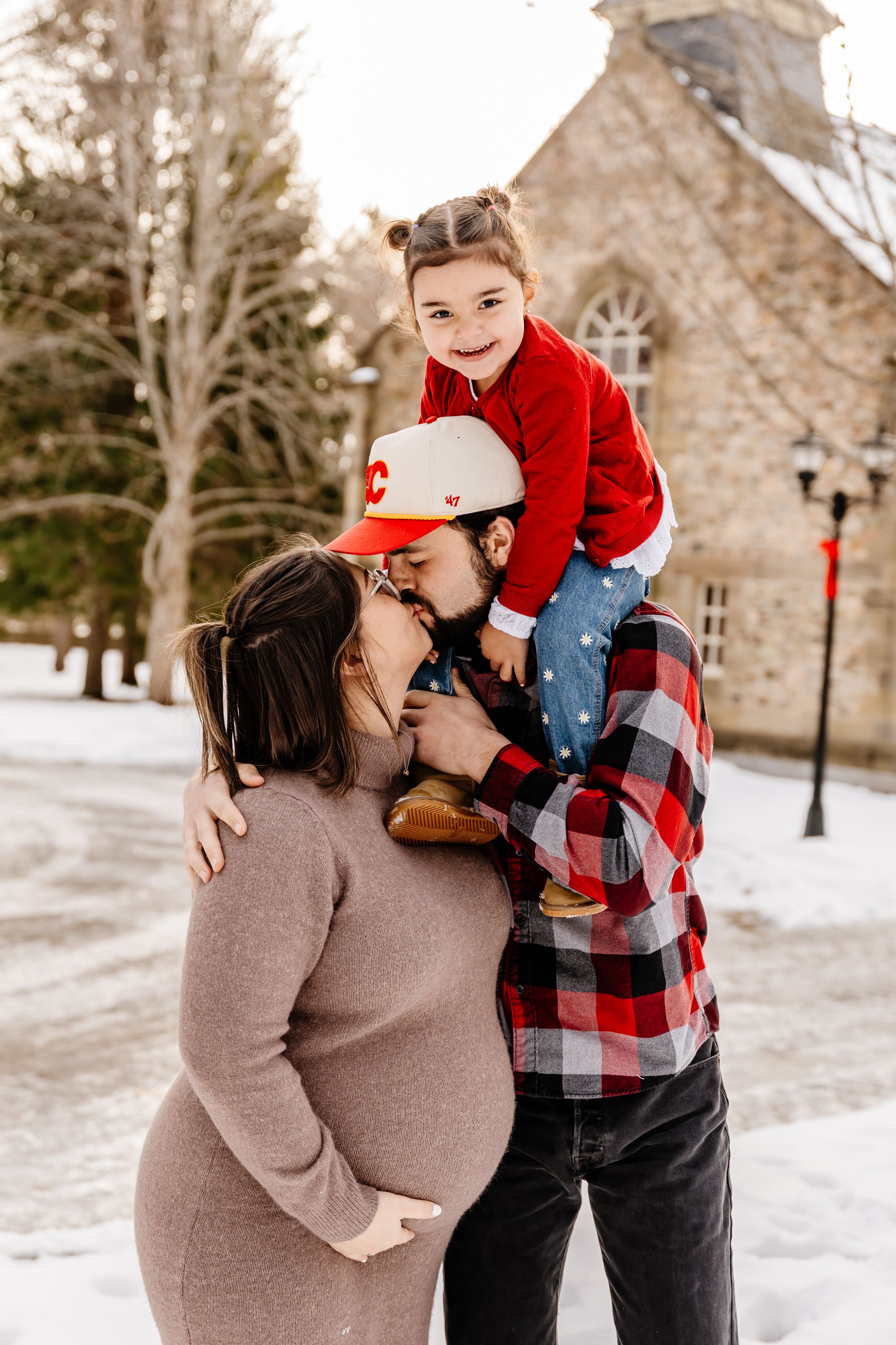Dad with toddler on shoulders kissing pregnant mom outside snowy stone building in winter.