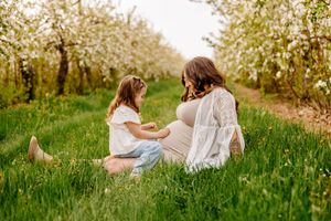 Pregnant woman sitting in blooming orchard with young daughter touching her belly in lush green grass.