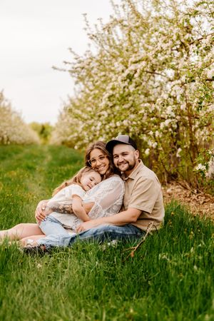 Happy family of three sitting on green grass near blooming apple trees in spring orchard.