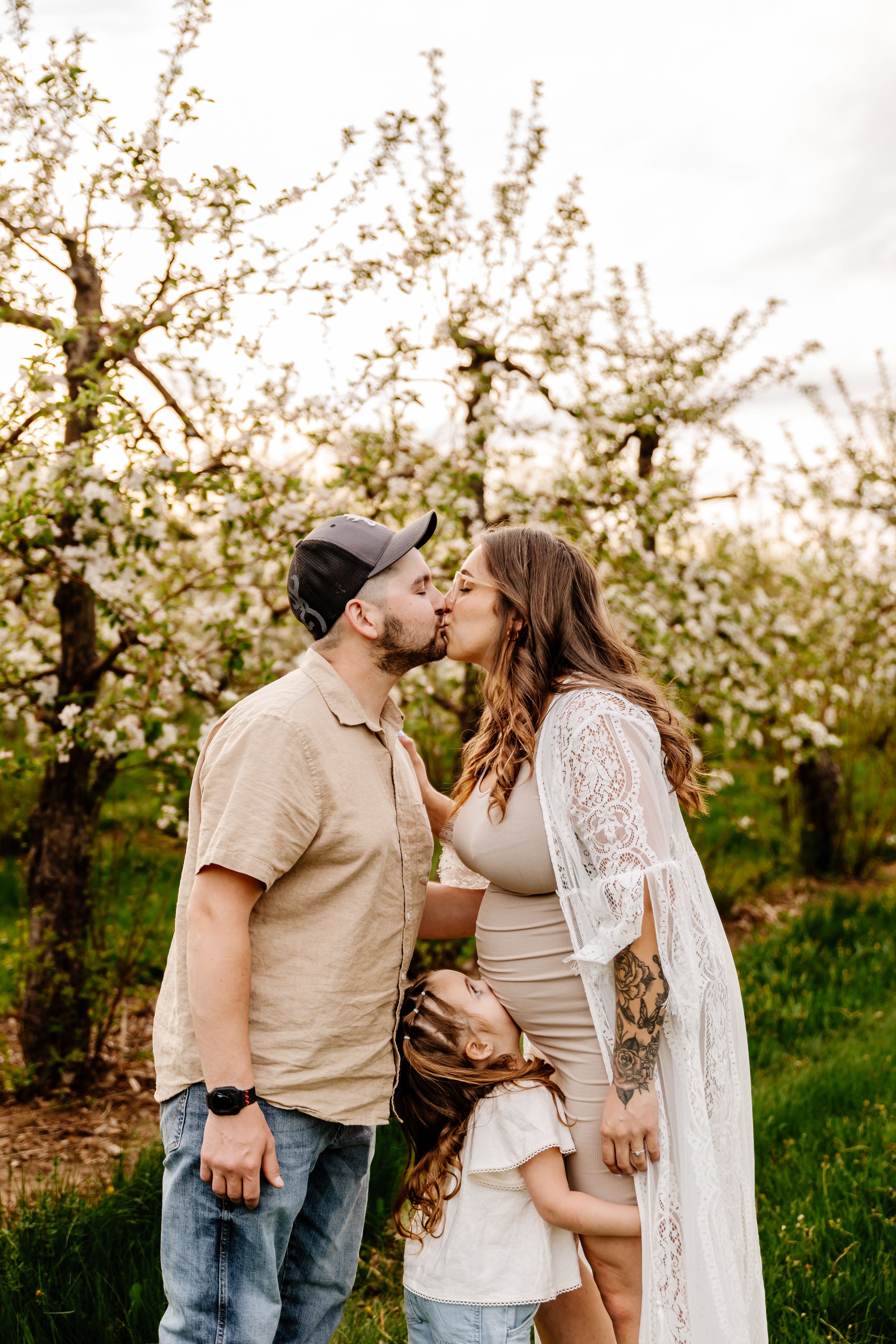 A couple kisses in a blossoming orchard while their young child stands between them in spring.